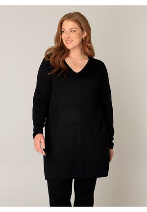 Lachende vrouw met lang rood haar draagt een zwarte tuniek met lange mouwen en zwarte broek, staand tegen een effen lichte achtergrond.