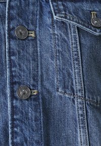 Blue denim fabric with a textured surface, featuring two metallic buttons and visible stitching. Pocket detail is prominent.