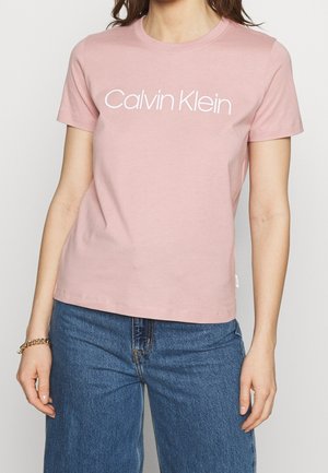 Femme portant un t-shirt rose Calvin Klein et un jean bleu, avec un bracelet en or au poignet gauche sur un fond neutre.