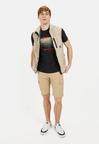 Beige letvægtsvest over en sort grafisk t-shirt, parret med khaki cargo shorts og hvide sneakers med marineblå detaljer.