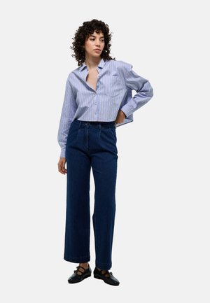 Chemise à rayures bleu clair avec une poche poitrine, assortie à un jean en denim foncé taille haute. Le modèle porte des chaussures noires.