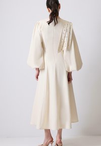 Egy nő hosszú, krémszínű ruhában áll, amely puffos ujjal rendelkezik, és a jobb vállán díszes drapéria feszül, mezítláb.