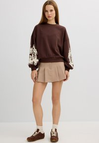 Mujer vestida con un sudadera marrón con mangas de encaje blanco, una falda mini plisada beige, calcetas blancas y zapatillas marrones de pie contra un fondo liso.
