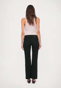 Mujer con pelo largo castaño, vistiendo una blusa sin mangas de color rosa claro, jeans negros de pierna recta y zapatos oscuros de slip-on, de pie y mirando hacia atrás.