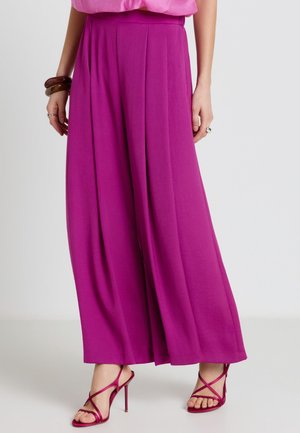 Femme portant un pantalon magenta taille haute à jambes larges, associé à des sandales à talons hauts à brides assorties et un haut rose.