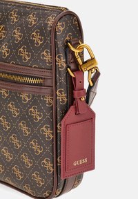 Brązowy wzorzysty róg torby Guess z złotym zamkiem błyskawicznym, złotym zapięciem i przymocowaną czerwoną skórzaną metką z napisem "GUESS" w złotych literach.