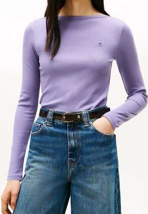 Kobieta ubrana w dopasowany liliowy, prążkowany, długorękawowy top, niebieskie dżinsy z wysokim stanem oraz ciemnobrązowy skórzany pasek z srebrną klamrą.