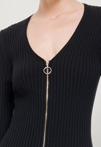 Musta ribbineuloksinen toppi, jossa syvä V-pääntie ja kultainen vetoketjusomiste, jossa on pyöreä vetolaite, vartaloa myötäilevä leikkaus ja teksturoitu pinta.