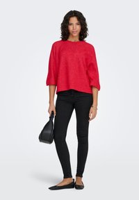 Pull rouge texturé et cintré avec manches trois-quarts ; associé à un jean skinny noir et des chaussures plates noires ; tenant un petit sac noir.