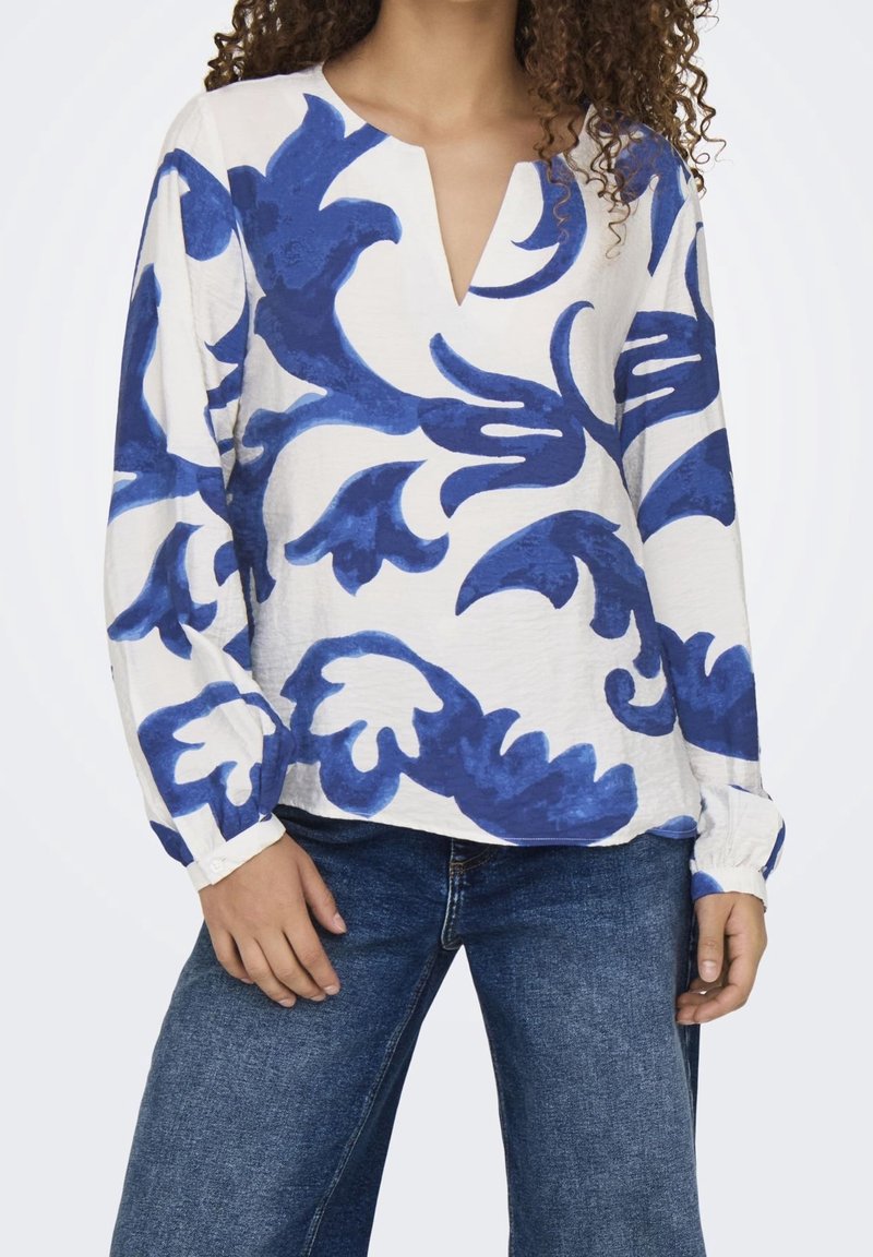 Blusa a maniche lunghe bianca con motivi floreali blu, dotata di scollatura a V e polsini arricciati. Il tessuto appare morbido con una leggera trama.