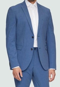 Mężczyzna ubrany w dopasowany niebieski garnitur z czarnymi guzikami, zestawiony z białą koszulą z guzikami, na jednolitym jasnym tle.