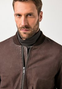 Uomo con capelli castani corti e barba, che indossa una giacca di pelle marrone scuro sopra un dolcevita nero, rivolto verso l'avanti su uno sfondo bianco.