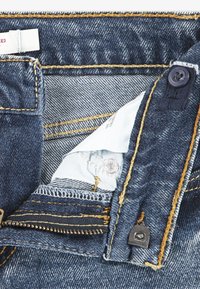 Jean en denim bleu foncé avec surpiqûres jaunes. Comprend un bouton en métal, une fermeture éclair et une étiquette d'entretien à l'intérieur de la ceinture. Détails du tissu texturé visibles.