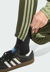 Černé tenisky s bílými pruhy, zelené sportovní kalhoty s světle zelenými postranními pruhy a zipem na kotníku, a černé krátké ponožky.