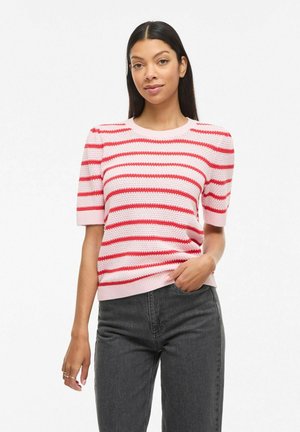 Frau mit langen dunklen Haaren, die einen kurzärmligen Pullover mit rosa und roten Streifen und schwarze Jeans trägt und mit einer Hand in der Tasche posiert.