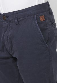 Marineblaue Hose aus strukturiertem Stoff, mit Seitentaschen, einem Knopfverschluss und einem braunen Lederlogo-Patch am Bund.