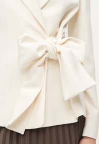 Κρεμ σακάκι με μεγάλο υφασμάτινο φιόγκο δεμένο στη μέση, σε συνδυασμό με καφέ υφασμάτινη φούστα.