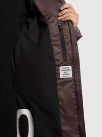 Brūnas izolējošas jakas iekšpuse ar melnu oderi, kas ietver rāvējslēdzēja kabatu un auduma etiķeti ar uzrakstu "Silts apskāviens no auduma."