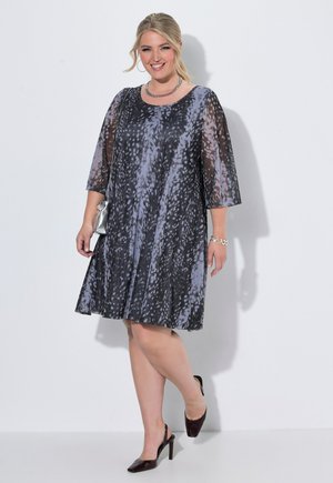 Donna sorridente che indossa un vestito nero e grigio a fantasia, lungo fino al ginocchio, con maniche trasparenti, tacchi neri, collana d'argento, braccialetto e piccola borsa argento.