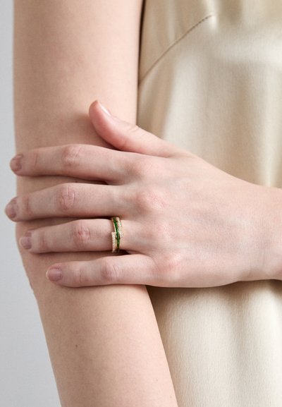 Anello d'oro con una banda di pietra verde e accenti trasparenti, poggiato su una mano contro uno sfondo di tessuto nudo. Texture liscia.