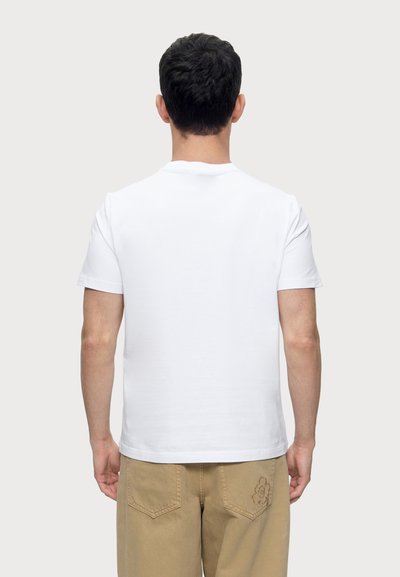 Homme de dos face à la caméra portant un t-shirt blanc uni à manches courtes et un pantalon marron clair avec un motif floral sur la poche arrière.