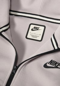 Lichtpaarse Nike zip-up jacket met zwarte rits, kraagafwerking met witte en zwarte strepen, en stoffen label met verzorgingsinstructies.