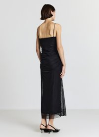 Schwarzes Midikleid mit dünnen Trägern, geraffter Textur am Rücken, transparenten Seitenteilen und geradem Saum. Kombiniert mit schwarzen Slingback-Pumps.
