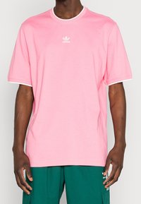 Uomo che indossa una semplice maglietta rosa di Adidas con il logo bianco sul petto e pantaloni verdi di Adidas con tasche laterali e strisce bianche.