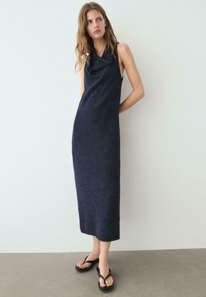 Kobieta o falowanych włosach nosi granatową, długą dzianinową sukienkę bez rękawów i czarne klapki, stoi oparta o gładką białą ścianę z rękami za plecami.