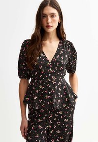 Blouse noire à motifs floraux avec manches bouffantes, décolleté en V et fermeture à boutons. Présente des motifs floraux roses et blancs sur un tissu texturé.