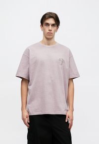 Φωτεινό μωβ oversized μπλουζάκι από μαλακό βαμβάκι, με στρογγυλή λαιμόκοψη και κοντά μανίκια. Περιλαμβάνει ένα μικρό κεντημένο λογότυπο στο στήθος.