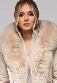 Beżowa pikowana kurtka z dużym kołnierzem z sztucznego futra. Kurtka ma przedni zamek błyskawiczny i gładką fakturę, łącząc ciepło i miękkość.