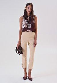 Blusa senza maniche a fiori scuri, pantaloni cropped beige chiaro con dettagli a bottone e accessori marroni. Tessuti lisci, design aderente.