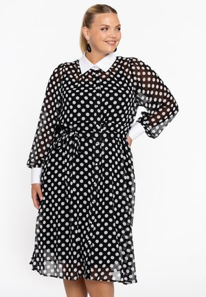 Robe noire à pois blancs avec un col blanc, des manches longues transparentes, une taille cintrée et une jupe fluide, ornée d'un motif de grands pois blancs.
