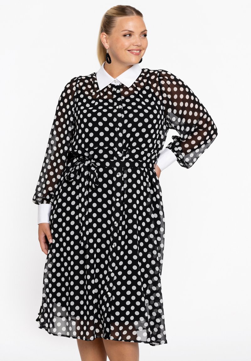 Schwarzes Kleid mit weißen Punkten, einem weißen Kragen, durchsichtigen langen Ärmeln, einer taillierten Taille und einem fließenden Rock, der mit großen weißen Punkten gemustert ist.