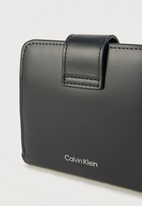 Μαύρο δερμάτινο πορτοφόλι με ομαλή υφή, διαθέτει κλείσιμο με καπάκι και ραμμένες λεπτομέρειες. Το λογότυπο "Calvin Klein" τυπωμένο σε λευκό χρώμα.