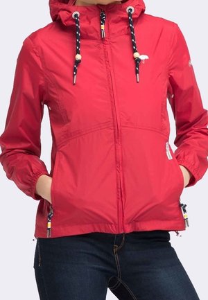 Czerwona kurtka z kapturem, czarno-białe sznurki, zamek błyskawiczny z przodu, elastyczne mankiety, boczne kieszenie, noszona z ciemnoniebieskimi dżinsami. Ręce w kieszeniach.