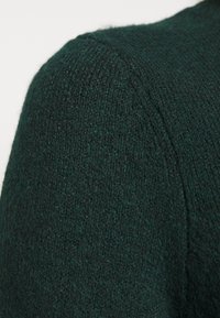 Épaule d'un pull en laine vert foncé montrant un tissu tricoté texturé et un détail de couture sur un fond clair uni.