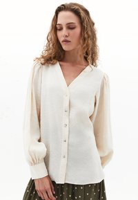 Crème blouse met een V-hals, knoopsluiting en pofmouwen. Zachte, gestructureerde stof met een subtiele glans en gerimpelde manchetten.