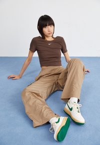 Brauner Cropped-Top mit kleinem weißen Logo, beige weite Hosen und weiße Sneaker mit grünen Akzenten auf einem blauen Hintergrund.