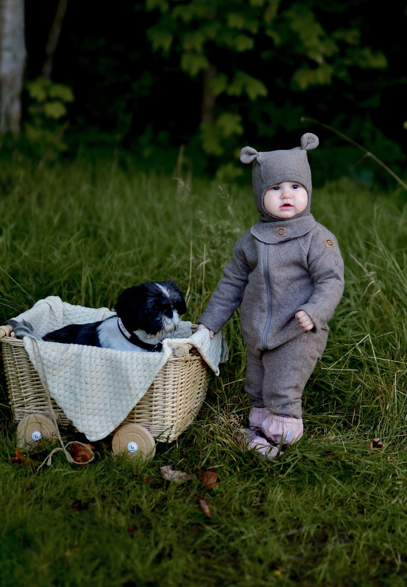 Hall hallast villast beebipükskostüüm karukõrvadega, esiküljel lukuga ja puidust nupuga. Lähedal korvis lebab must-valge koer.