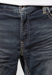 Niebieskie dżinsy o wyblakłym wykończeniu i kontrastowych żółtych szwach, z guzikiem zamiast zamka oraz przednimi kieszeniami.