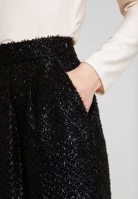 Jupe noire texturée avec une surface duveteuse, dotée de poches latérales et d'une taille haute. Le tissu réfléchit la lumière de manière subtile.