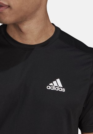 Erwachsener trägt ein schwarzes Adidas-T-Shirt mit weißem Adidas-Logo auf der linken Brust, leicht nach links blickend, vor einem einfarbigen Hintergrund.