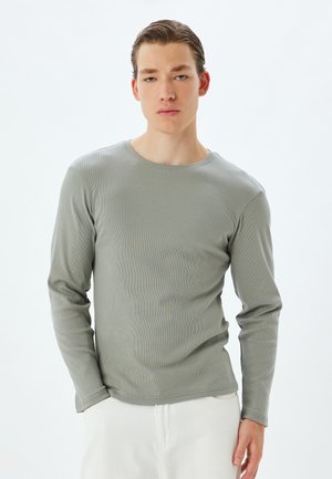 Camiseta de manga larga gris de canalé con cuello redondo, diseño ajustado y textura suave, combinada con pantalones de color claro.