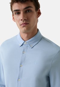 Lichtblauw polo shirt met een button-down kraag, twee zichtbare knopen en een subtiele reliëf "B" logo op de linkerborst. Gladde textuur.