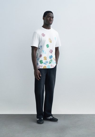 Ενήλικος άνδρας που στέκεται, φοράει λευκό μπλουζάκι με πολύχρωμα σύμβολα δολαρίων και διαμάντια, μαύρο παντελόνι και μαύρα αθλητικά παπούτσια με λευκές ρίγες.