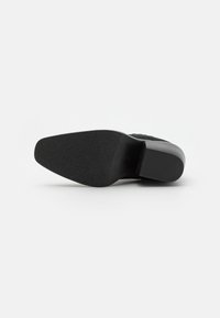 Chaussure noire à bout pointu vue par dessous, montrant une semelle texturée et un petit talon bloc sur fond blanc.
