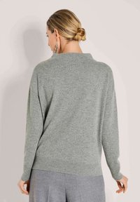Frau mit blonden Haaren zu einem Dutt getragen, die silberne Ohrringe und einen hellgrauen langärmeligen Pullover sowie lockere graue Hosen trägt, mit dem Rücken zur Kamera gewandt.