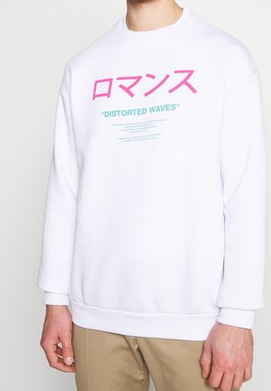 Hvid langærmet sweatshirt med lyserød japansk tekst, turkis "DISTORTED WAVES" og mindre turkis beskrivende tekst båret af person i beige bukser.